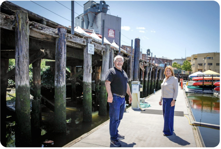 Screen shot from headline image in Petaluma Voice article. Mayor Kevin McDonnell and member Rosemary Hart are standing on the docks in front of the trestle. The Great Petaluma Mill is in the background.